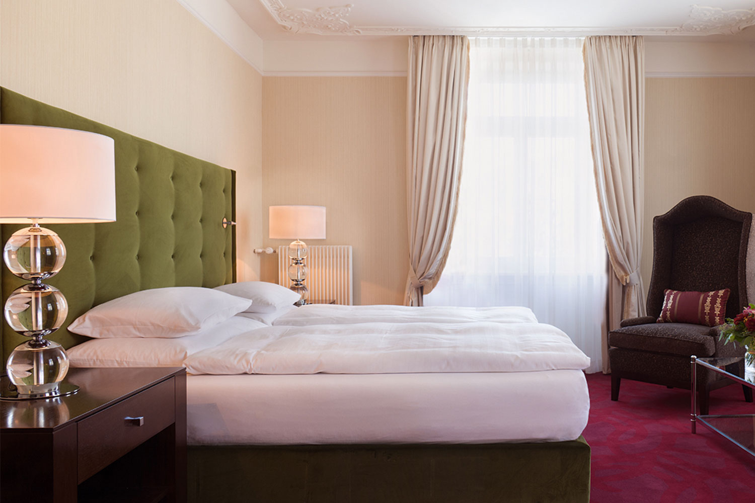 An interior shot of a one bed suite. The bed is donned in white linens and the headboard is a dark green. The carpet is red and the two high back chairs in each corner of the room are brown. There are two bedside tables with lamps on top of them.