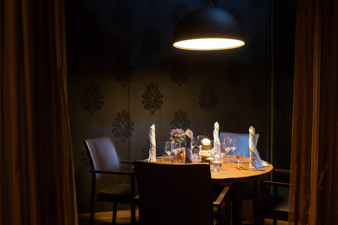 an interior shot of a private dining area. There is a small round wooden table set with intricately folded napkins and a purple flower as the centerpiece. There are four brown buttery leather chairs around the table. There is a light above the table and the wallpaper is a dark gray with a Victorian pattern.