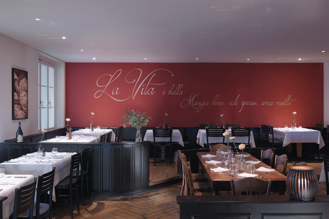The dining room of Pomodoro. The wall is a deep red color with white cursive writing. The tables are donned in white linens with a single pink rose as the centerpiece of each table. The tables are lined with black chairs.