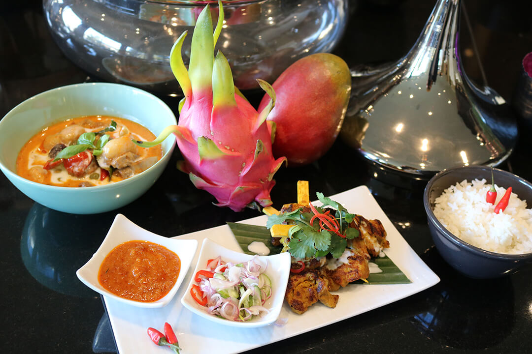 A detail shot of a diner order. There is a bowl of spicy soup with colorful garnishes in a light blue bowl. A bowl of white rice with red pepper garnish in a gray bowl. And a long white plate with meat and little bowls of sauce and garnishes. The is a dragon fruit and a mango on the table for decoration