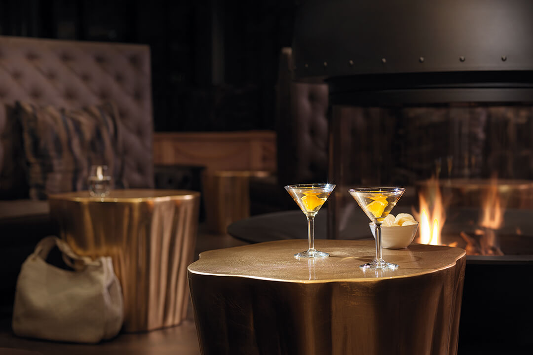 an interior shot of the summit bar. There is a gray fireplace in the center of the room, Surrounding the fireplace are brown leather couches.. Next to each couch are two dark brown leather chairs and a gold table. On one of the gold tables sits two martini glasses with golden liquid in them.