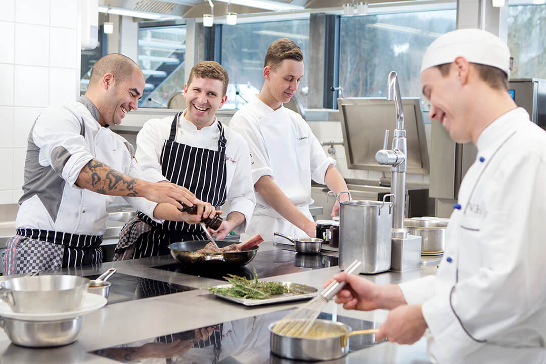 a group of chefs in white uniforms stand around a counter. Each are attending to their own dishes. They hold whisks and spatulas. The surrounding kitchen is stainless steel and impeccable organized.