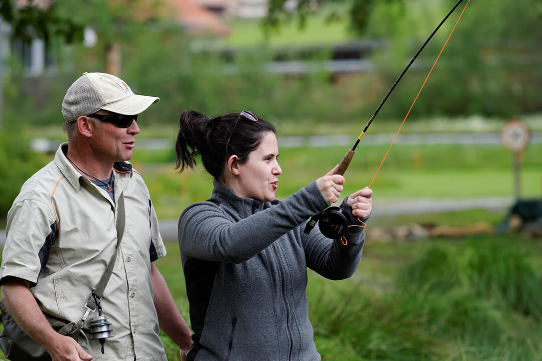 a man in a pale green hiking button up shirt and cap stands behind a woman with a gray fleece jacket and her hair tied in a bun. She is holding a large fishing rod in front of her. They are both looking out into the distance.