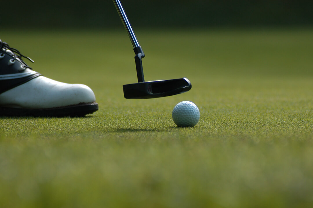 detail shot of a white golf ball perched on the green lawn. A putter hovers over the ball and there is a black and white golf show visible in the corner of the photo