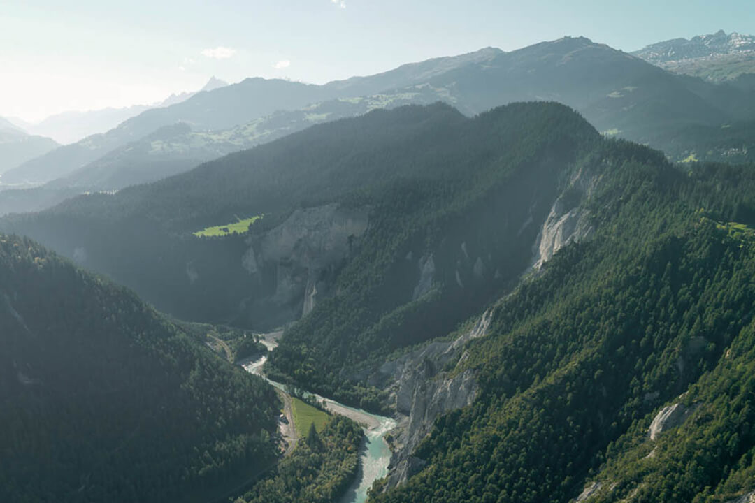 an ariel shot of a bright blue river that snakes its way through the valley between two mountains. The mountains are covered in evergreen trees. The sun shines down and reflects off the river.