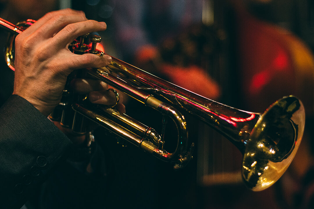 a close up shot of a musician in a gray suit playing the trumpet. The trumpet is sleek and gold. The musicians hands are placed gently on the keys of the instrument. There is a red light in the background