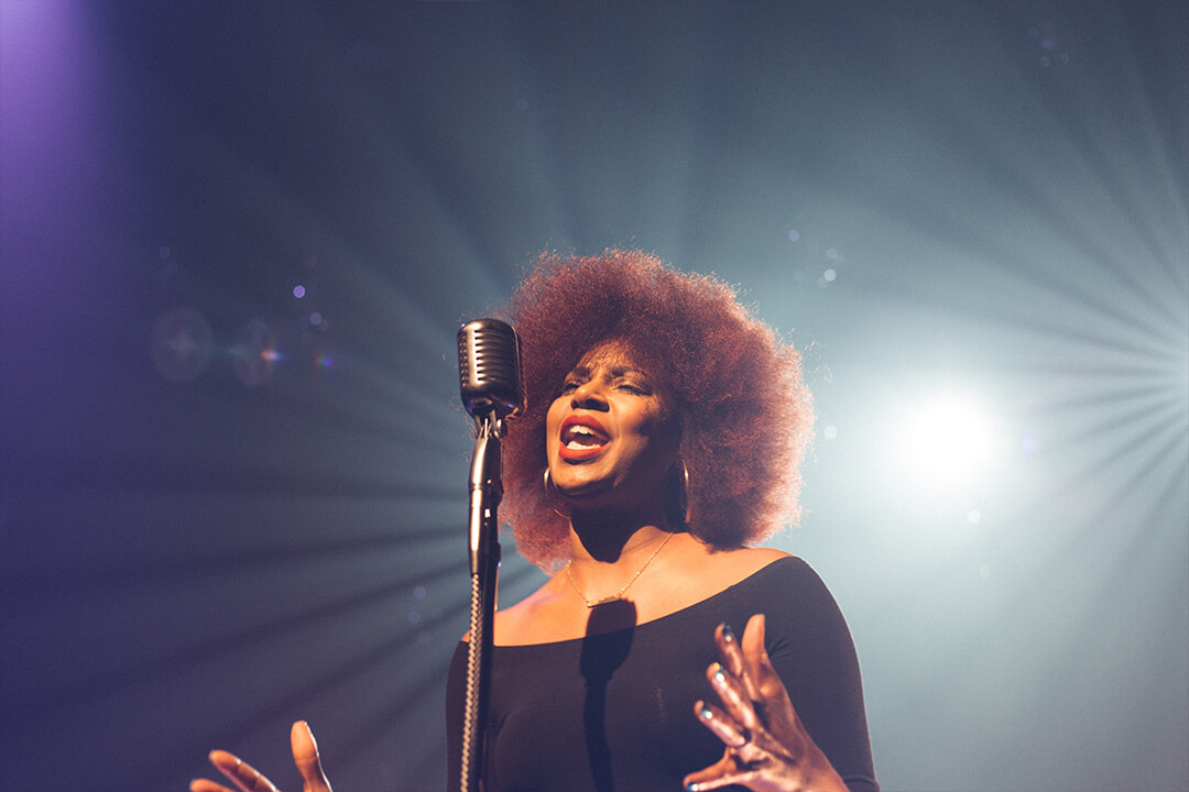 a woman in a black off the shoulder top and red lipstick sings into a vintage microphone. Her hands are outstretched and her face is lit with emotion. Behind her is a bright spotlight.