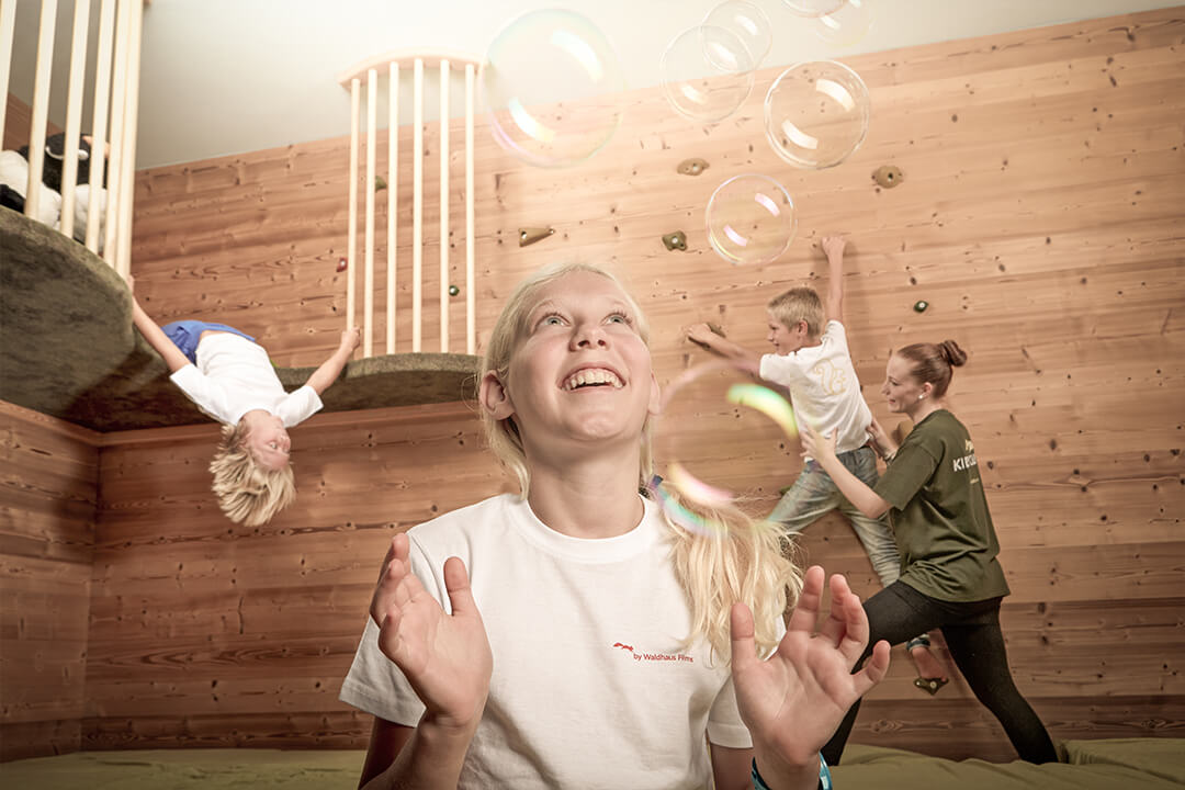 a young girl with a long blond pony tail wearing a white shirt looks up in wonder at the bubbles dropping down in front of her. In the background two boys and an instructor are playing on a rock wall.
