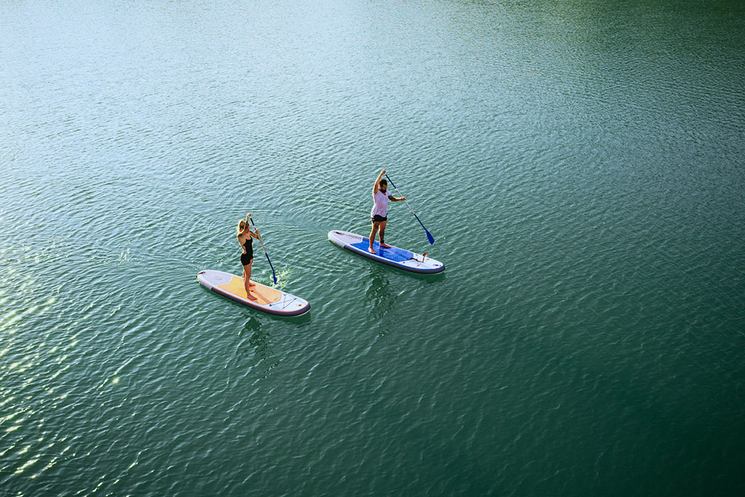 a man and a woman stand on paddle boards. The man is riding on a blue paddle board and the woman is riding on an orange paddle board. They are using paddles to push themselves through the turquoise waters of Lake Cauma