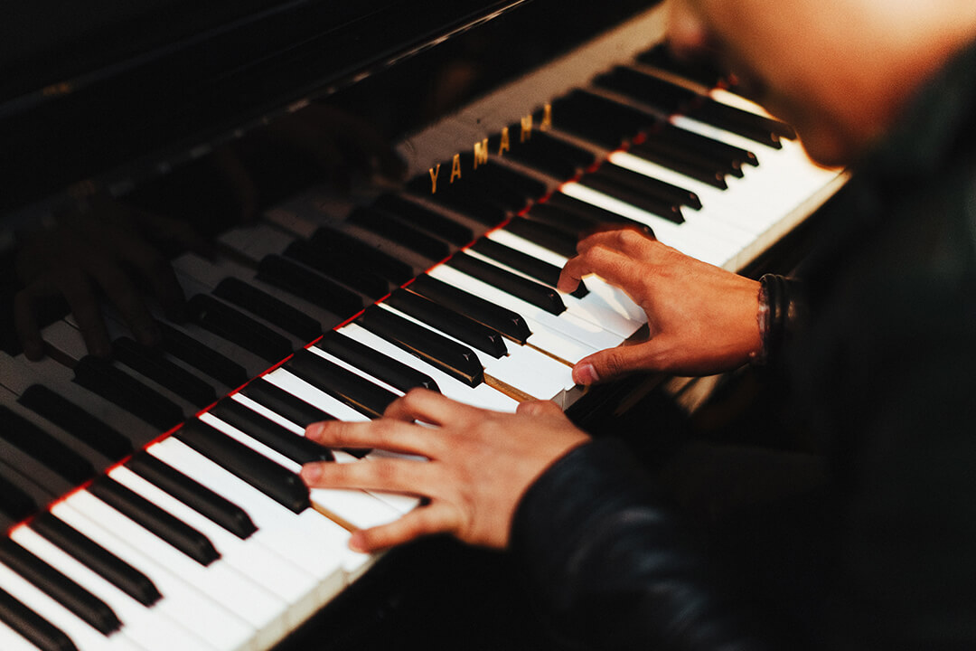 a man is sitting hunched over a piano. His fingers are extended to reach multiple keys. The piano is a sleek black.