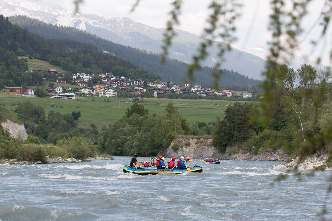 three large blue and yellow rafts paddle down a choppy river. In the distance is lush green farmland and a tiny village at the base of the swiss alps.