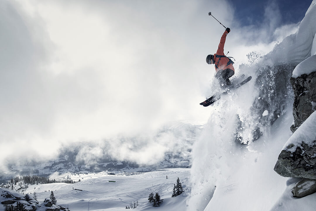 a skier in a red jacket flies through the air. He has skiied of of a drop off and the snow sprays around him. In the background are evergreen trees and the swiss alps. A large white cloud obstructs the full view of the mountain range.