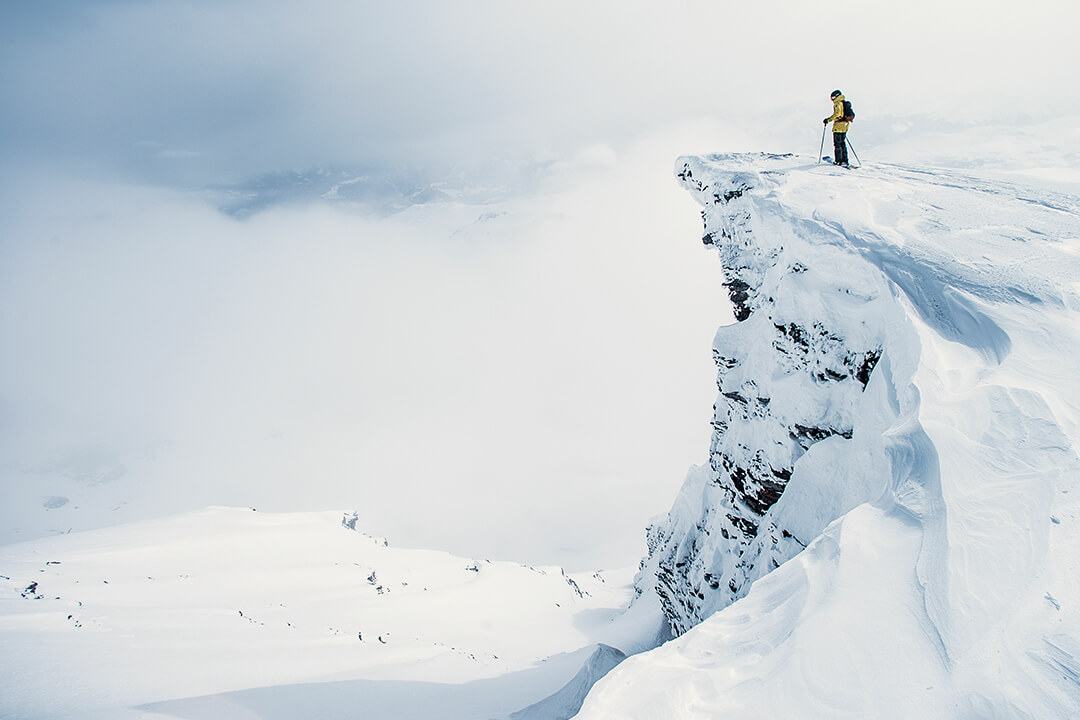 a skier in a bright yellow jacket looks out over a cliff. The background of the photo is covered by white clouds.