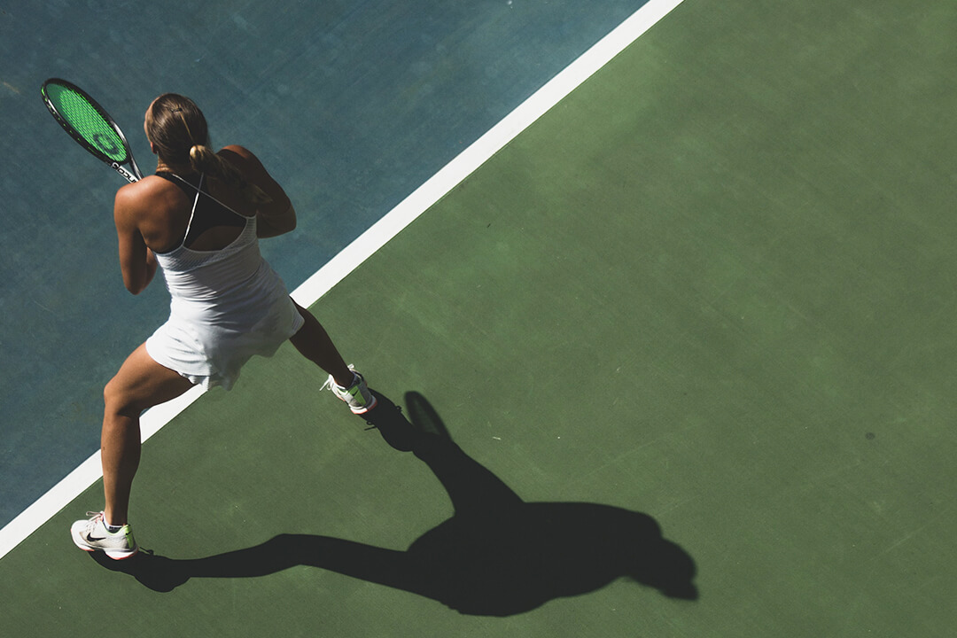 a woman in a white tennis dress and white tennis shoes stands in a crouches position as she waits for the ball to be served. Her hair is tied back in a tight ponytail and she hold her neon green racket close to her chest