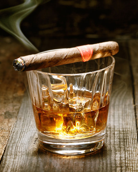 detail shot of a glass cup with amber colored whiskey in it with two ice cubes. Resting on top of the glass is a lit cigar. The smoke from the cigar slowly winds its way up into the air.
