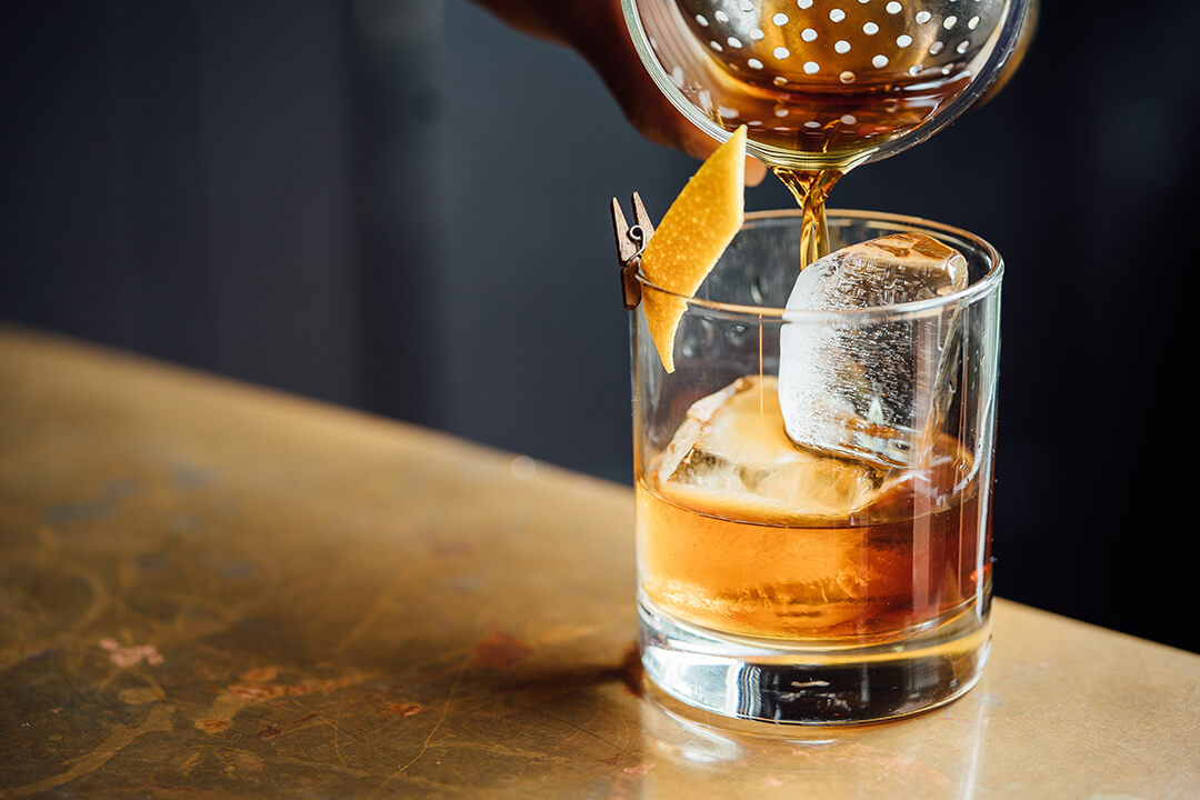 a detail shot of a mixer pouting a whiskey cocktail into a glass cup with large ice cubes. There is a little orange garnish clipped to the side of the cup. The glass rests on a worn gold table.