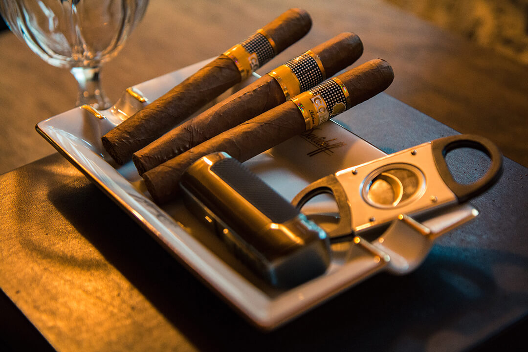 a detail shot of of a white platter with three cigars with gold wrapping, a silver lighter and a silver and black bottle opener. In the background is the shape of a glass cup.