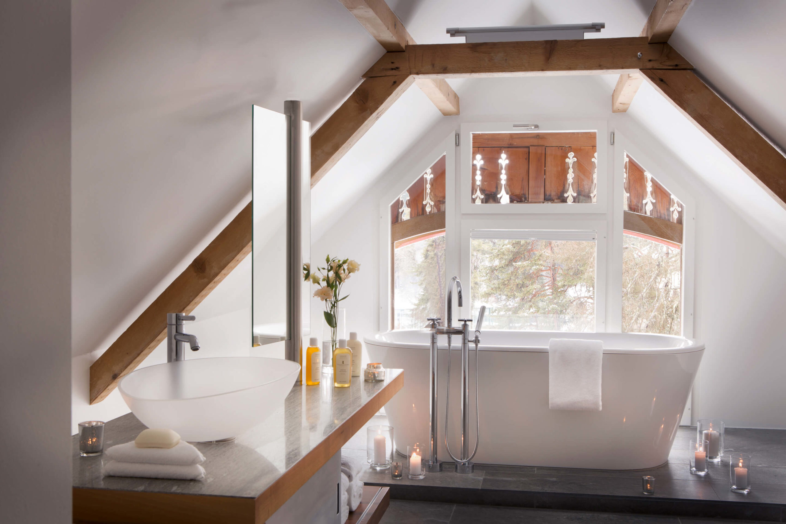 charming bathroom, white bath tub with window view of the trees, vanity mirror, sink, towels, beauty products, candles, flowers