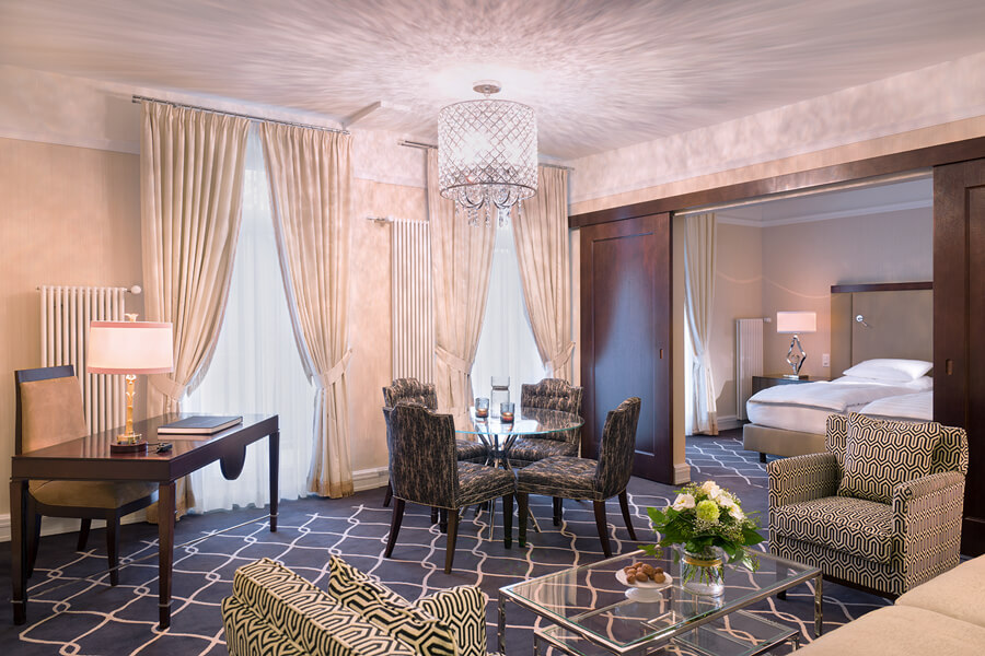 suite with white bed, sofa, chairs, dining table, dining chairs, desk, chairs, lamps, and french doors open to balcony
