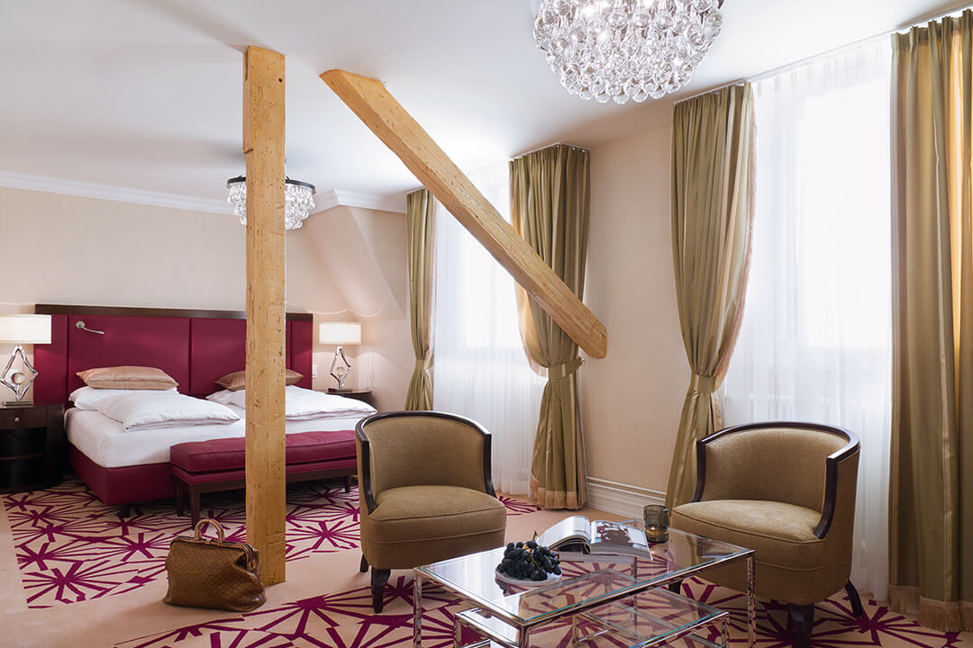 interior shot of the superior junior suite, The bed is donned in white linen with a burgundy headboard and bench. There are green curtains and a red and white patterned rug. There are two neutral chairs around a glass coffee table. Wooden beams break up the space.