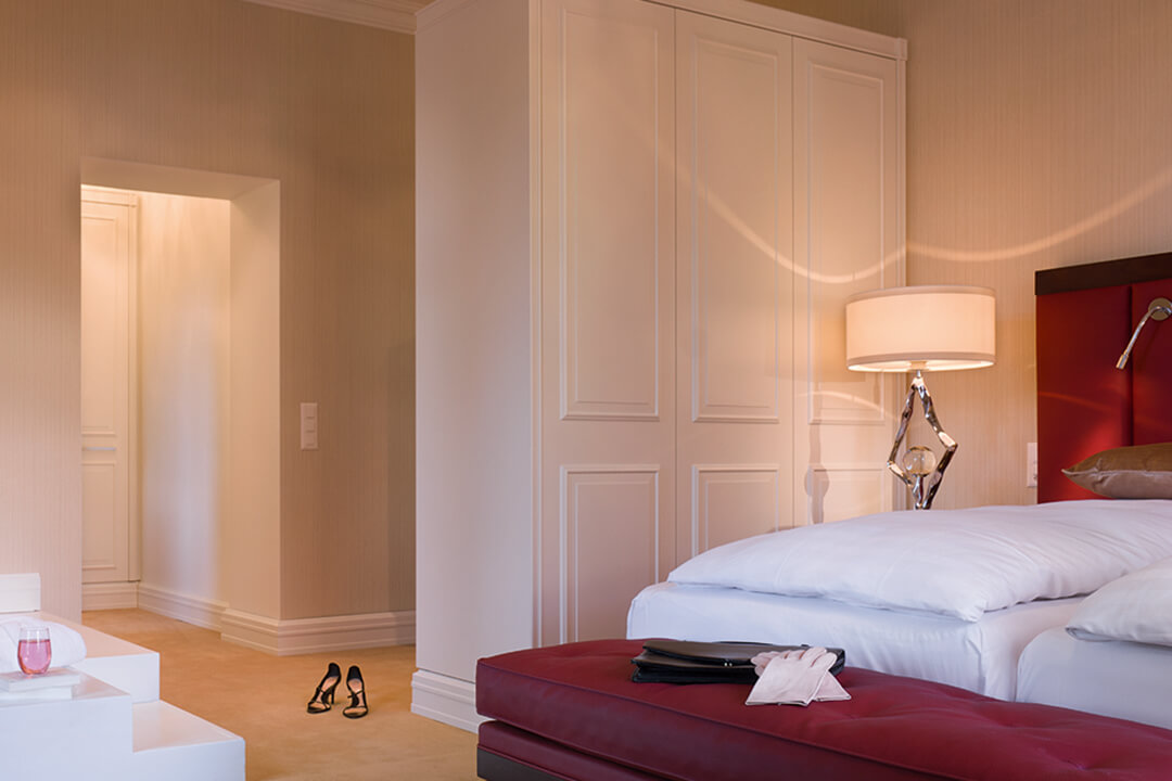 an interior shot of a two bed suite. The two beds are donned in white linens and there are accents of red around the room in the headboards, bench, and carpet. There is a white stand alone tub in the corner of the room.