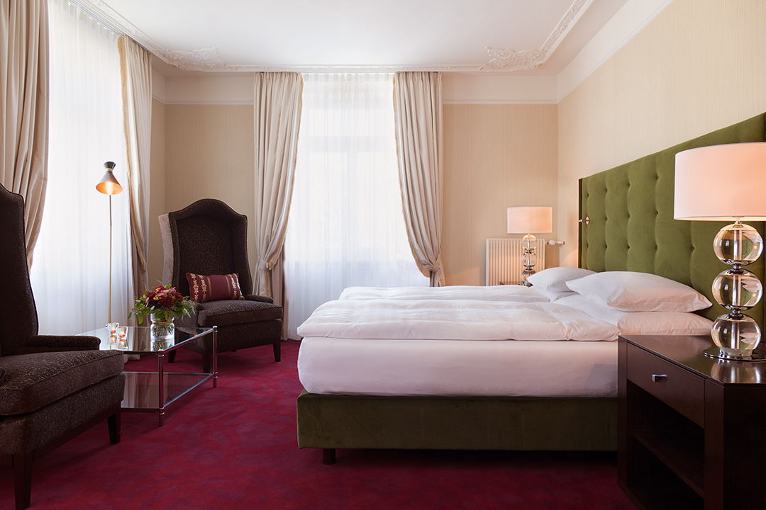 An interior shot of a one bed suite. The bed is donned in white linens and the headboard is a dark green. The carpet is red and the two high back chairs in each corner of the room are brown. There are two bedside tables with lamps on top of them.