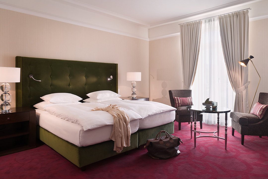 An interior shot of a one bed suite. The bed is donned in white linens and the headboard is a dark green. The carpet is red and the two high back chairs in each corner of the room are brown. There are two bedside tables with lamps on top of them.