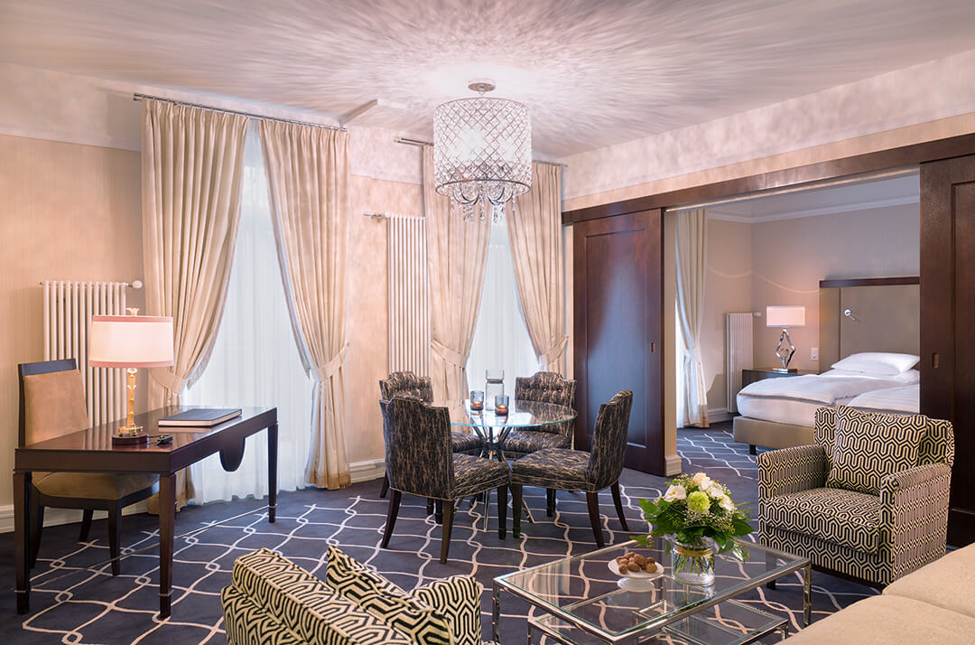 suite with white bed, sofa, chairs, dining table, dining chairs, desk, chairs, lamps, and french doors open to balcony