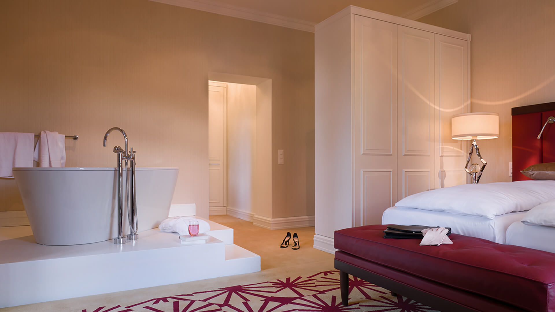 an interior shot of a two bed suite. The two beds are donned in white linens and there are accents of red around the room in the headboards, bench, and carpet. There is a white stand alone tub in the corner of the room.