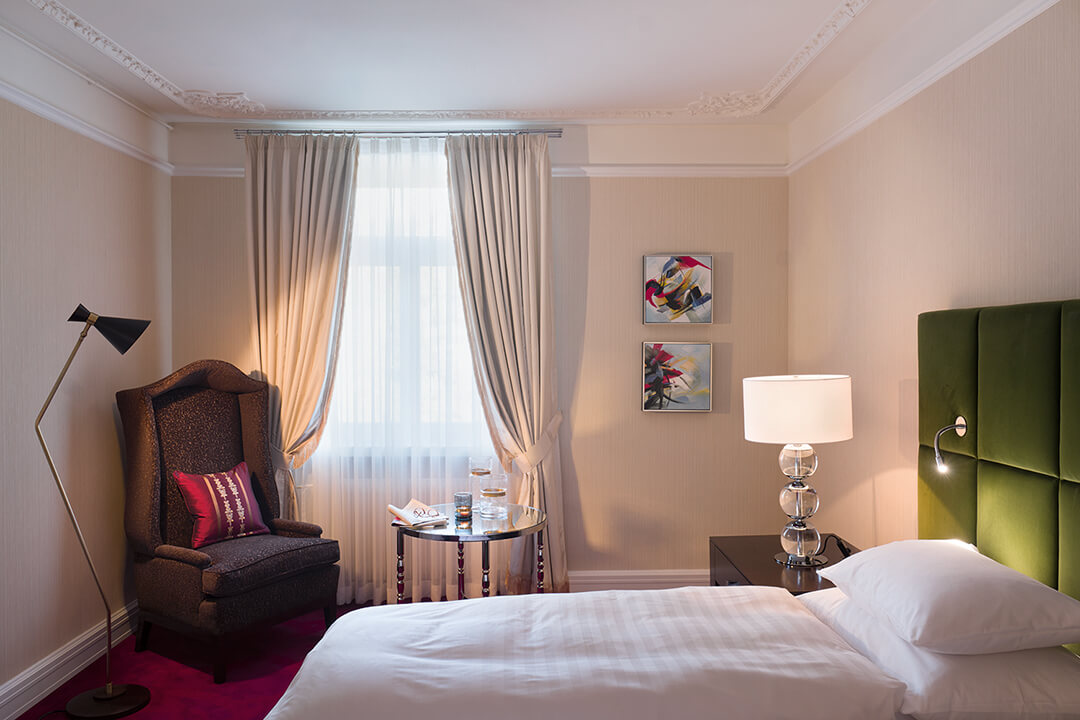 an interior shot of a one bed suite. The bed is donned in white linens and the headboard is a forest green. There is a large brown chair in the corner of the room next to a window. There is a small round table with a candle, a book and reading glasses on top of it.