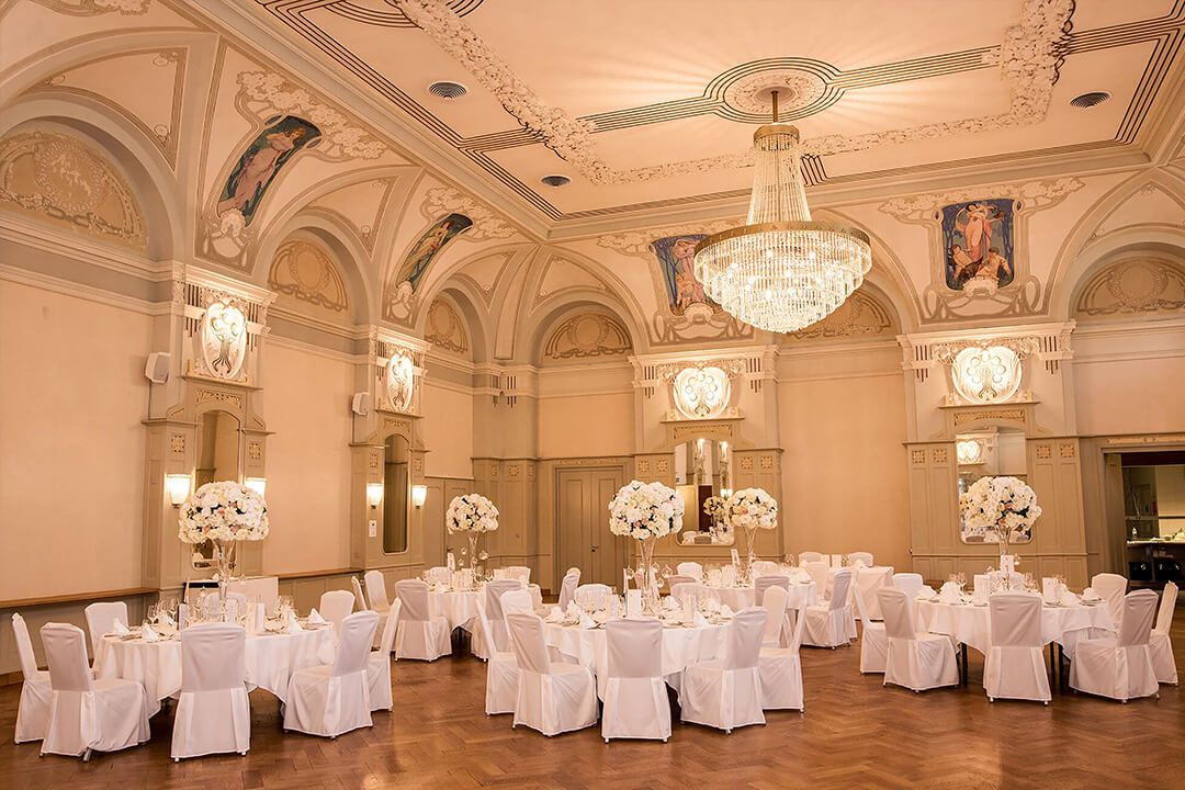 An interior shot of the Belle Epoque Hall decorated for a reception. There are many round tables donned in white linens, elegantly set with large white and green floral centerpieces. There are white chairs circling the tables. Hanging from the ceiling are three large crystal chandeliers.