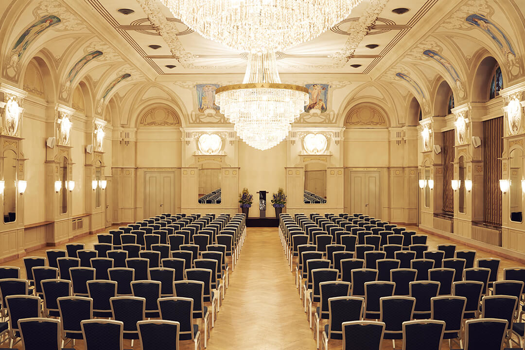 an interior shot of Belle Epoque Hall decorated for a speech. There are many rows of navy chairs and a podium at the front.
