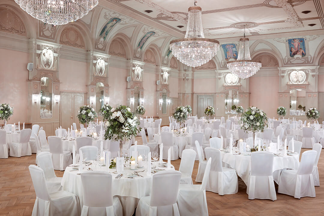 An interior shot of the Belle Epoque Hall decorated for a reception. There are many round tables donned in white linens, elegantly set with large white and green floral centerpieces. There are white chairs circling the tables. Hanging from the ceiling are three large crystal chandeliers.