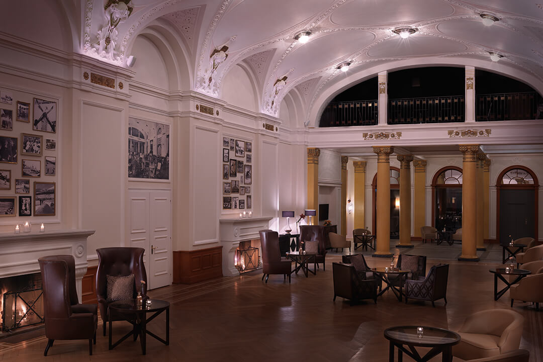 An interior shot of the Belle Epoque Pavilion. At the far end of the room are golden pillars. There are circular brown tables around the room with brown leather chairs with pillows circling each table. There is a fireplace on the left wall. Along the wall are vintage photographs of scenes from the resort.