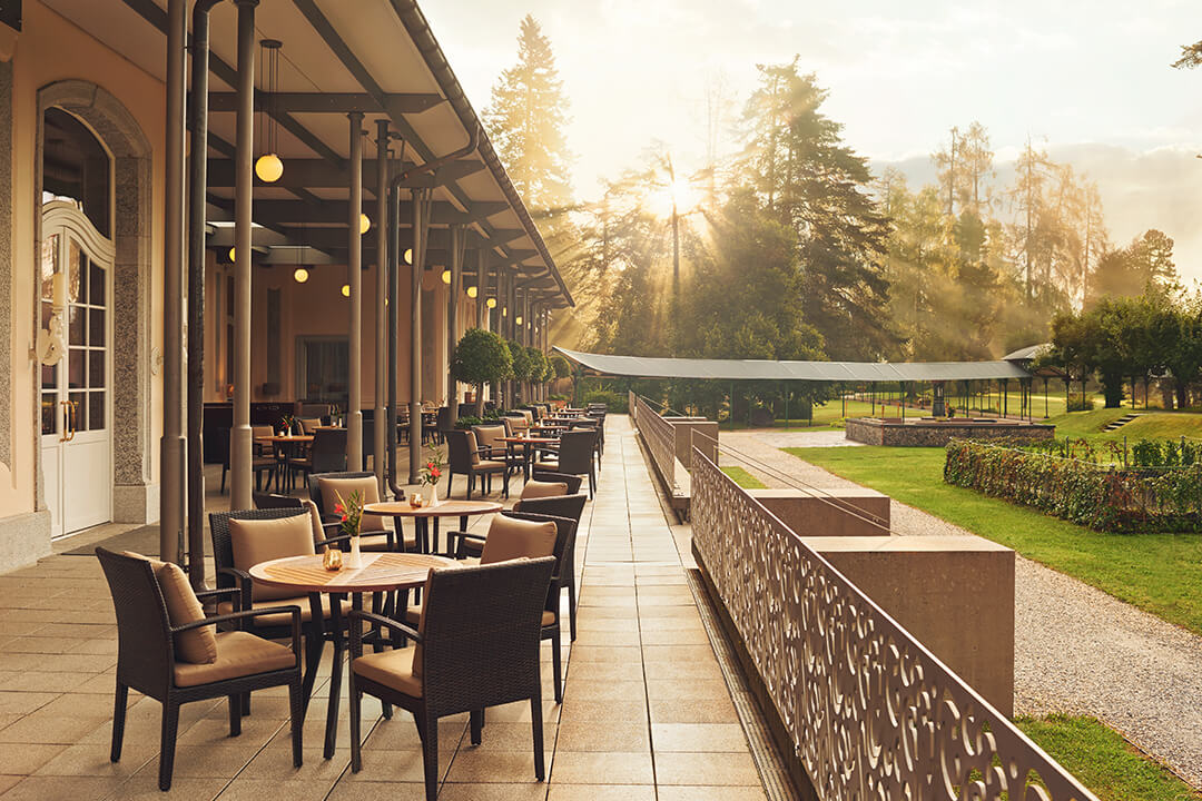 exterior shot of an outdoor dining patio. the patio is lined with black woven chairs with tan cushions surrounding light brown round table tops. There is a candle and a floral arrangement at each table. The patio overlooks the lush green grounds. In the background there are large evergreen trees with the sun shining through.