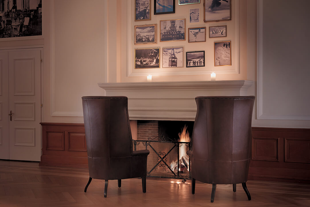 An interior shot of the Belle Epoque Paviliion. Two high back brown leather chairs sit facing a lit fire. Above the fireplace are vintage photos of skiers and ice skaters.