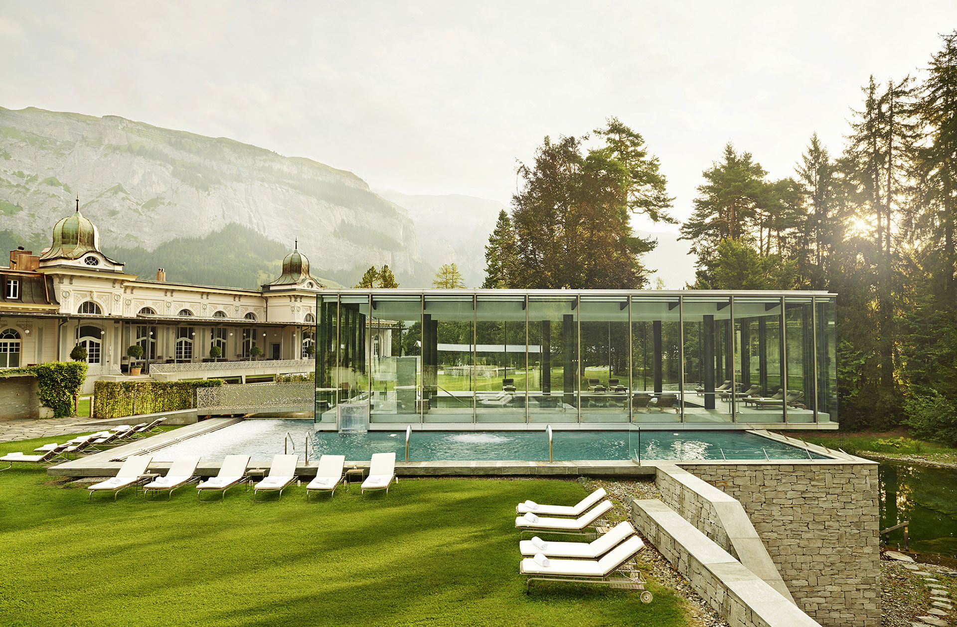 exterior shot of the waldhaus flims spa building and pool. The building is white with decorative fixtures at the top. The pool is indoor and outdoor. The indoor portion of the pool is encased in a glass fixture. The outside portion of the pool is lined with white lounge chairs. In the distance are evergreen trees and the swiss alps.