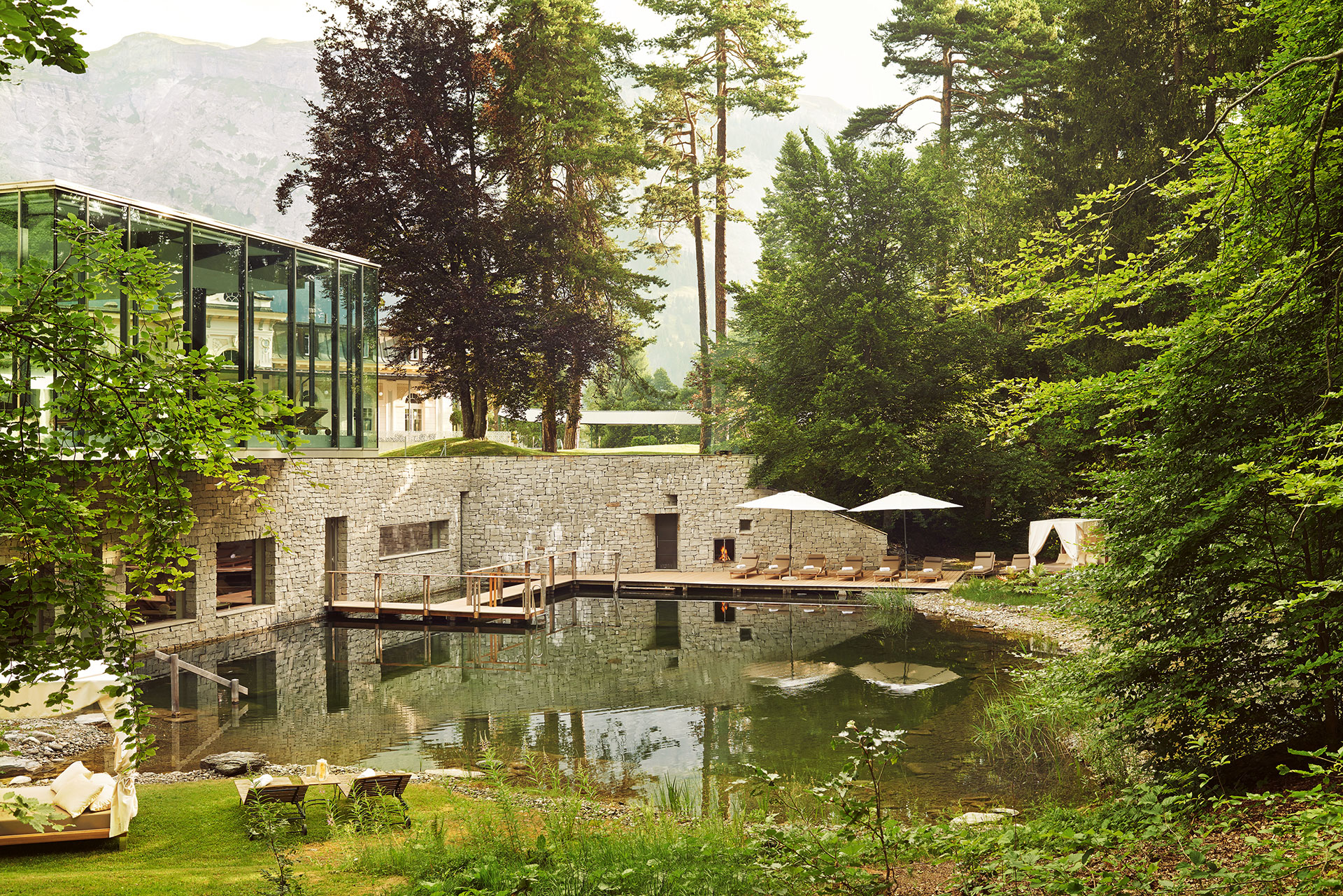 Landscape shot of the resort's pool. The pool is surrounded by lush greenery. The pool leads into the spa and there is a wooden bridge that leads guests inside to the outside and vice versa. Lining the pool are brown lounge chairs and white umbrellas. In the background there is a view of the swiss alps