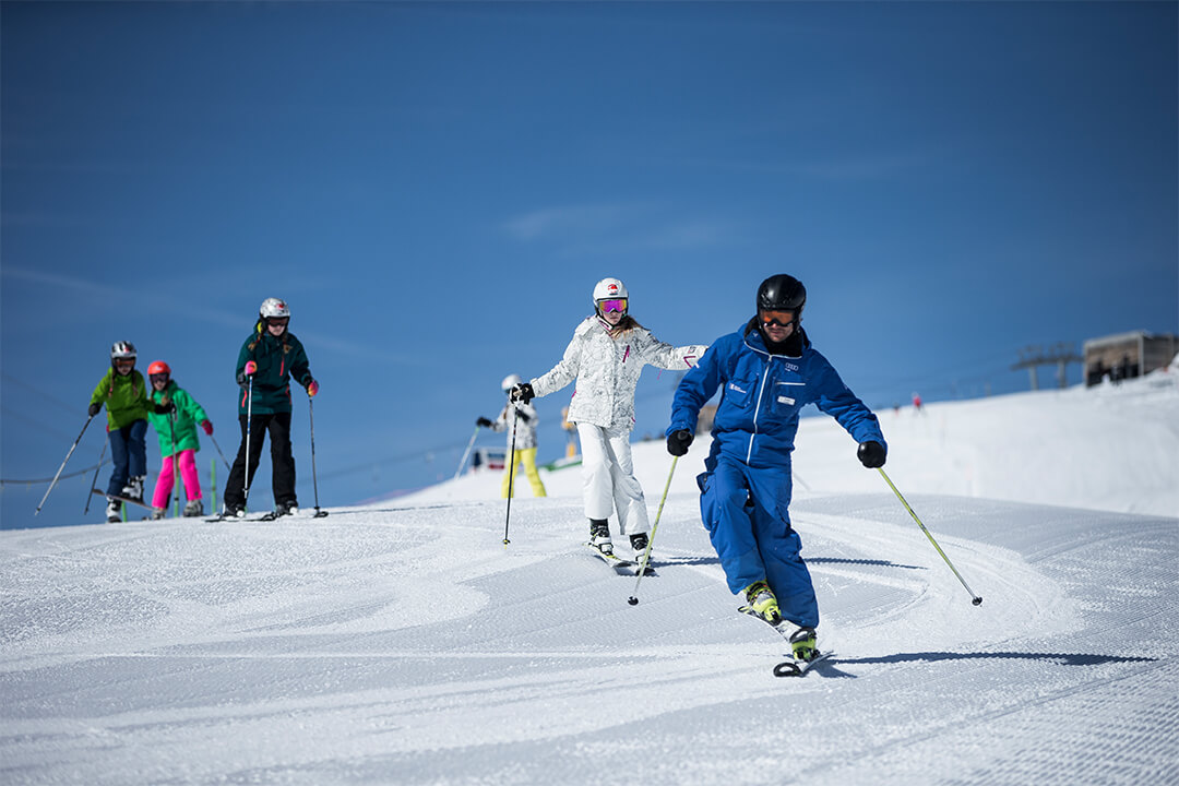 a ski instructor in an all royal blue outfit leads a line of children down a well groomed slope. The sky in the background is a deep, clear blue.