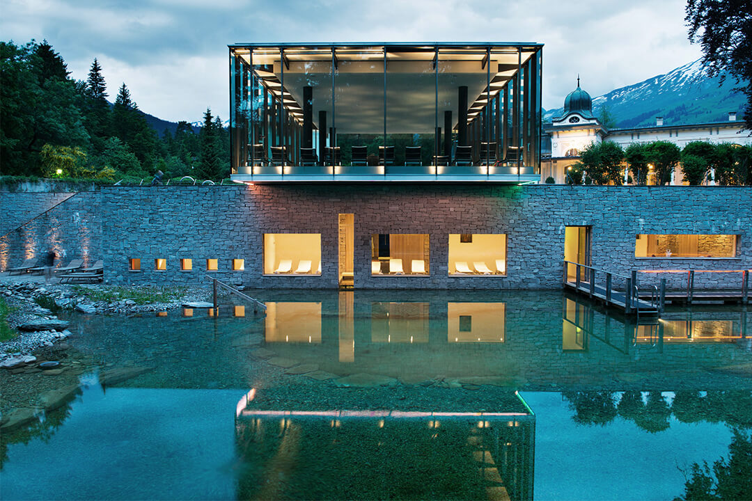 an exterior shot of the resort's spa and outdoor pool. The building is modern and layered. The pool is natural, blending seamlessly into the surrounding nature. There are golden lights coming from the windows of the spa and it is reflecting off of the pool. In the distance are the evergreen trees and tops of the swiss alps.