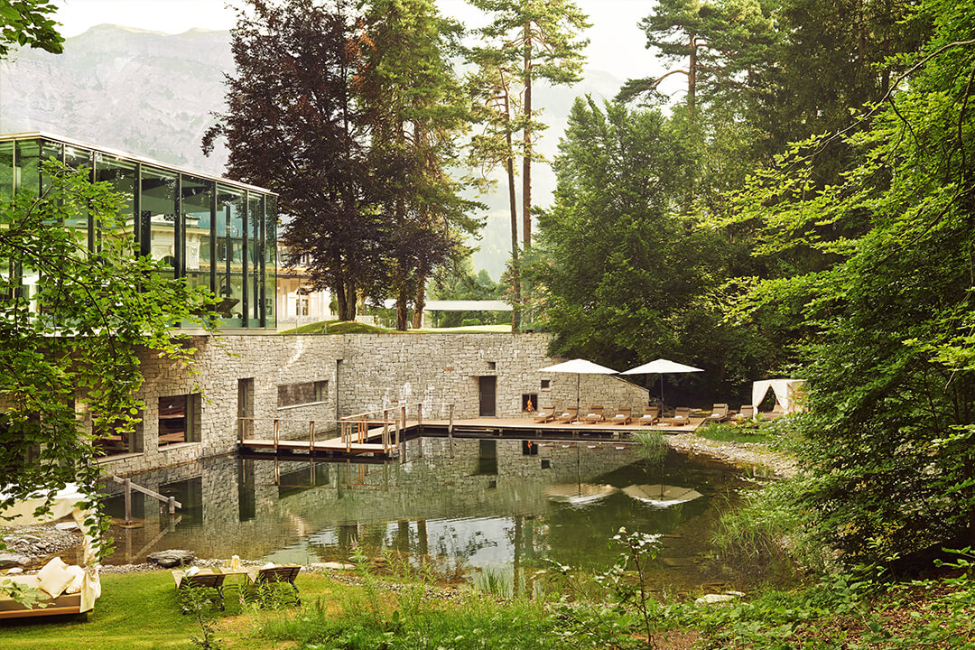 Landscape shot of the resort's pool. The pool is surrounded by lush greenery. The pool leads into the spa and there is a wooden bridge that leads guests inside to the outside and vice versa. Lining the pool are brown lounge chairs and white umbrellas. In the background there is a view of the swiss alps