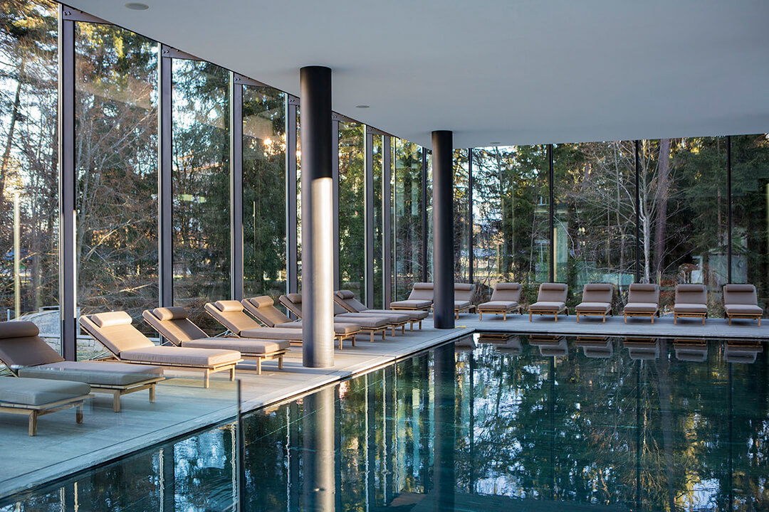 interior shot of the resort's indoor pool. The pool is a deep blue color and reflects the gray lounge chairs that line it. There are black pillars around the room. The walls are floor to ceiling windows giving a view of the forest surrounding the resort.