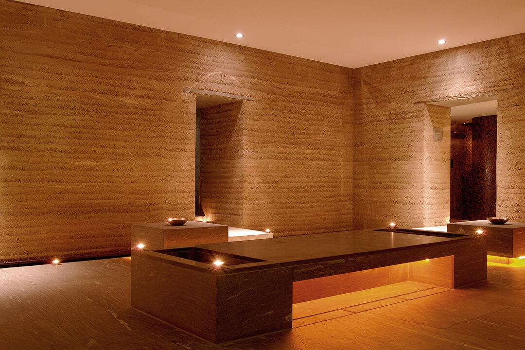 a golden tinted room with a long bench in the center of it. he walls are textured and light brown. There are lights that give the room a golden glow.