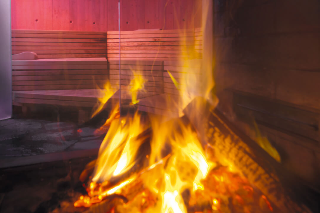 a fire blazes in the foreground of the photo. In the background you can see through glass floor to ceiling windows a sauna. The sauna is tinted red and there are multiple levels of light wood benches.
