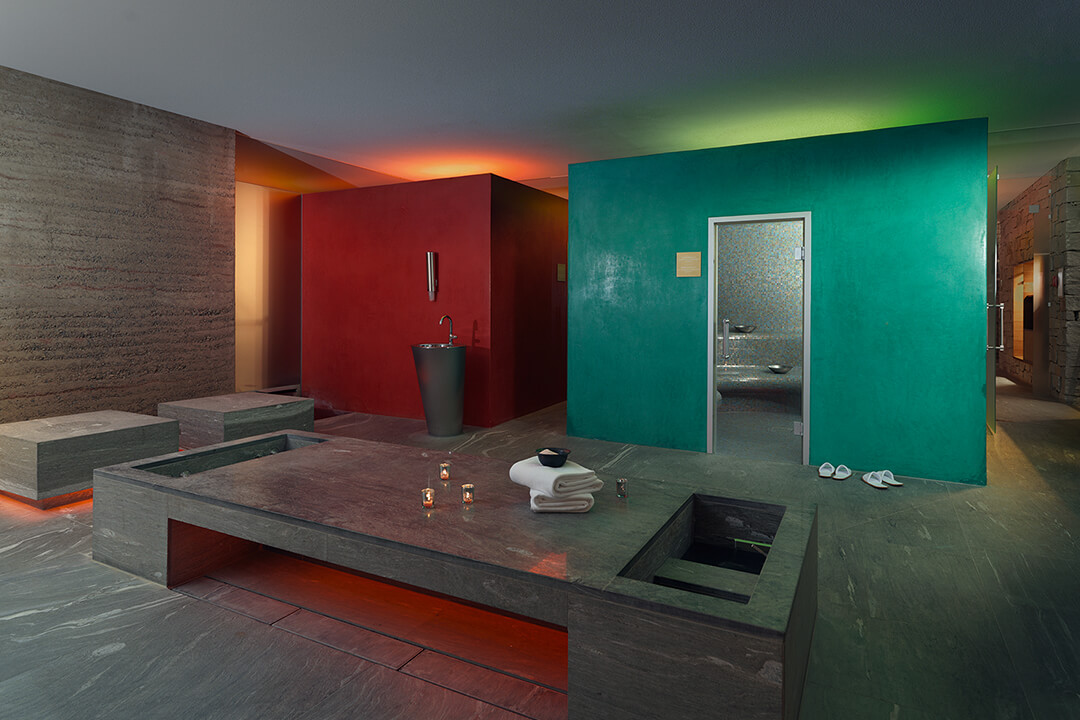 interior shot of a sauna. The walls are reds and blue and the floors are a slate gray. There is a long table in the middle of the room with towels and candles resting on it.