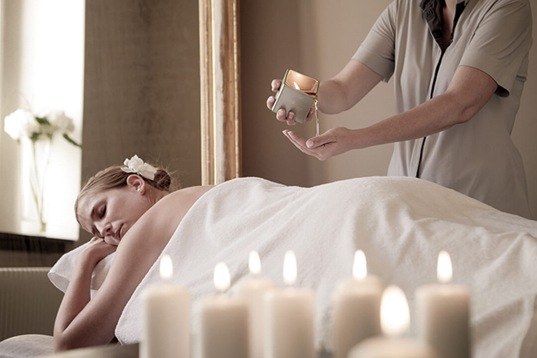 a woman lies on her stomach on a massage table. Her face is tilted toward the camera, her eyes are closed and she looks relaxed. Another woman in a gray uniform stands over her and pours a liquid on her hands to massage into the woman's back