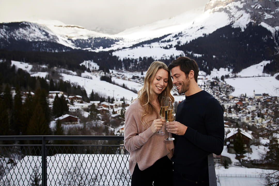 the couple is at the rooftop terrance, they are being close together and cheering with 2 glasses of champagne, the background is the snow cover town, trees and mountains